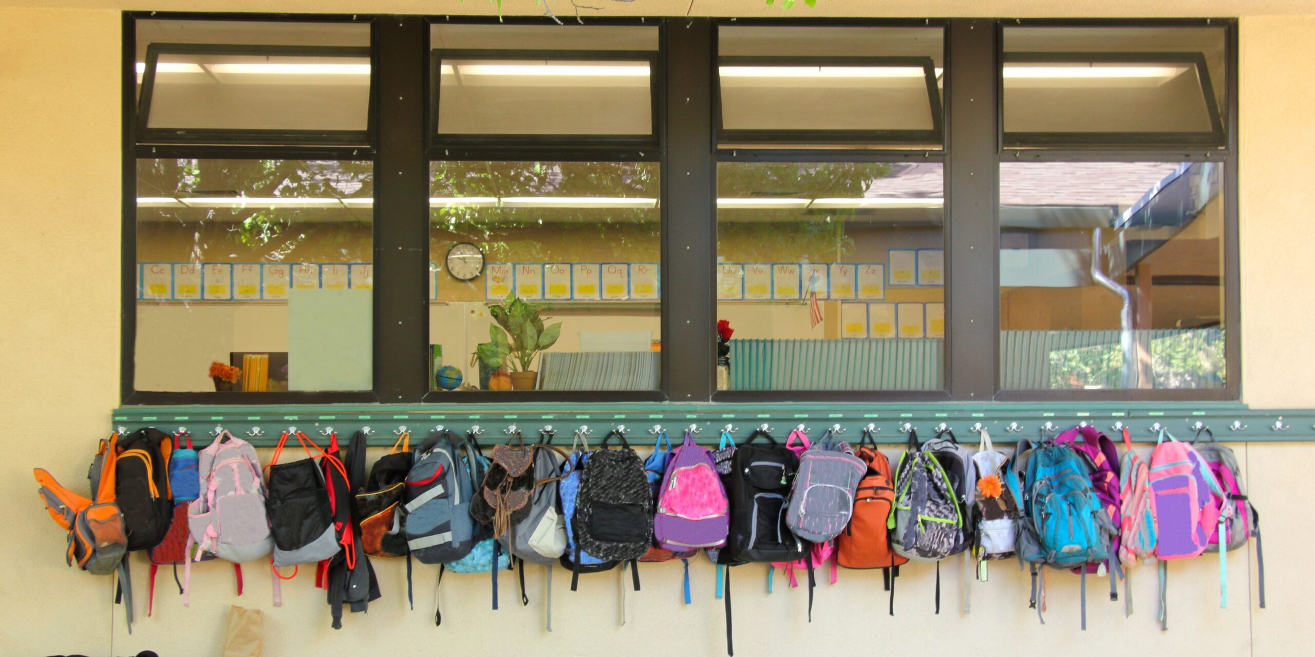 A line of colorful backpacks hung up outside of a school window.