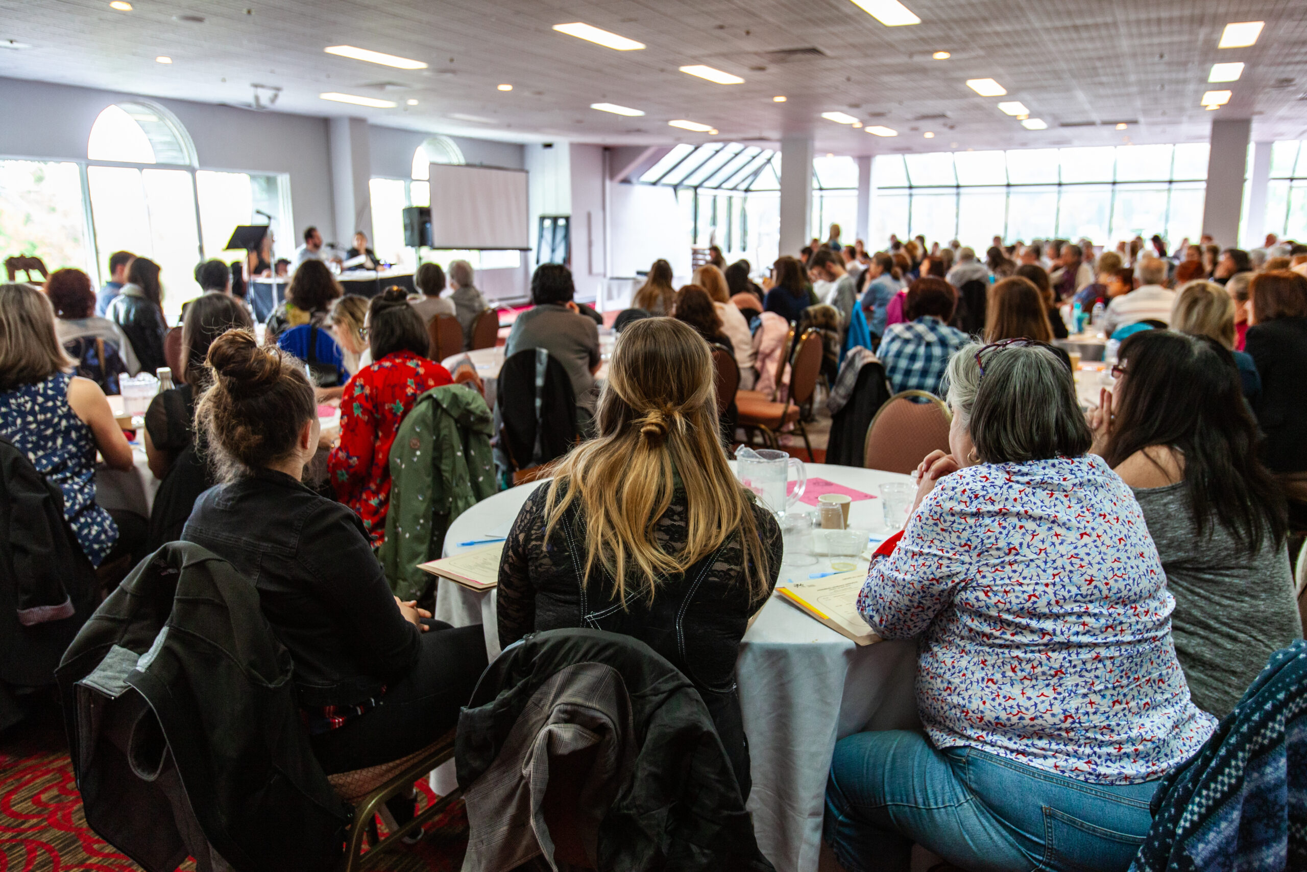 A back view on a group of women sitting at a large table during a networking event for white collar workers. Lots of people can be seen watching a presentation